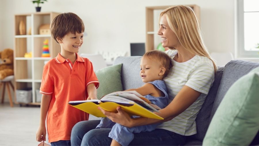 Blond woman with a small child sitting on her lap reading a book and smiling to older child.